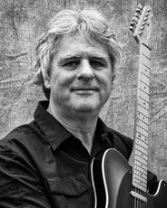 Person holding a guitar, smiling at the camera against a textured outdoor wall.