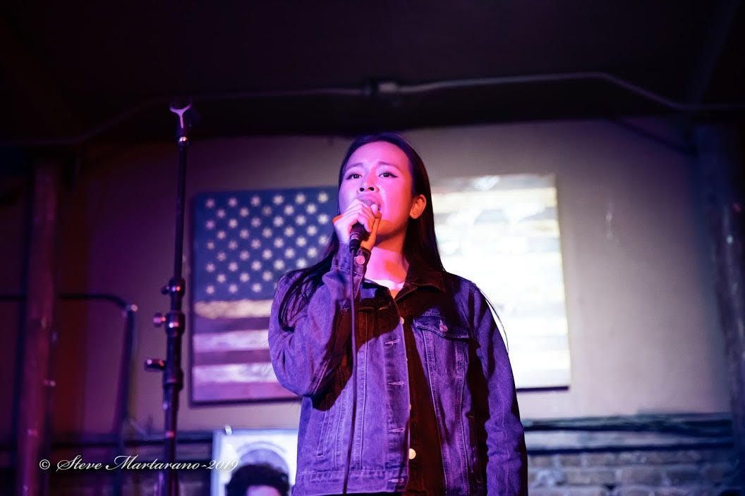 Singer performing on stage under purple lights, holding a microphone with an American flag backdrop.
