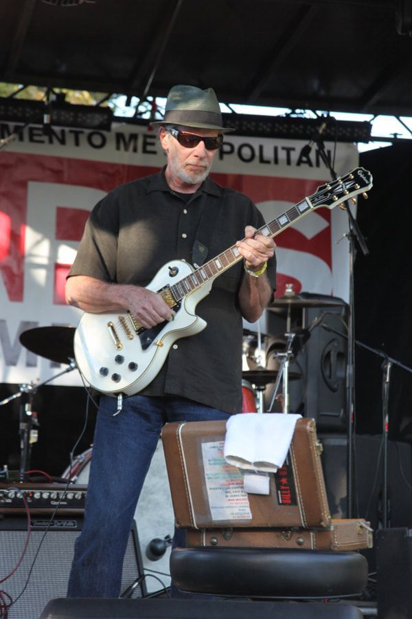 Musician in black shirt and hat playing a white electric guitar on an outdoor stage