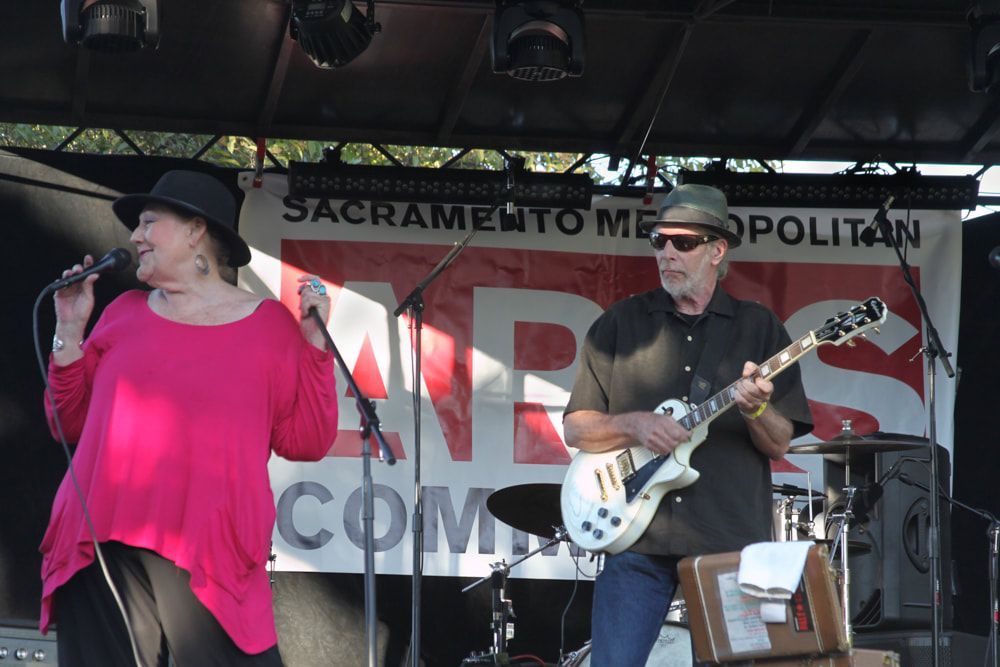 Singer in bright pink top and guitarist in black perform on an outdoor stage under a Sacramento banner.