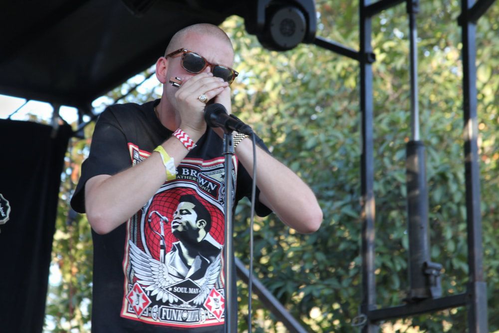 Singer performing into a microphone outdoors, wearing sunglasses and a black graphic T-shirt.