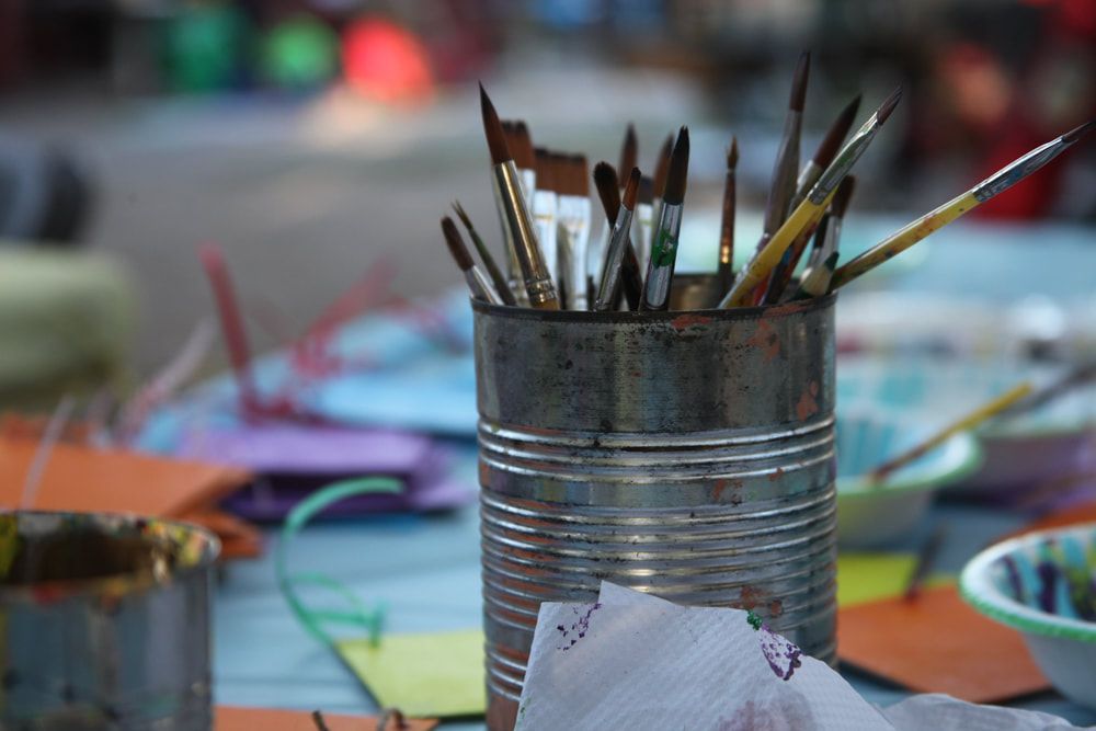 Paintbrushes in a tin can on a colorful art table, with blurred supplies in the background