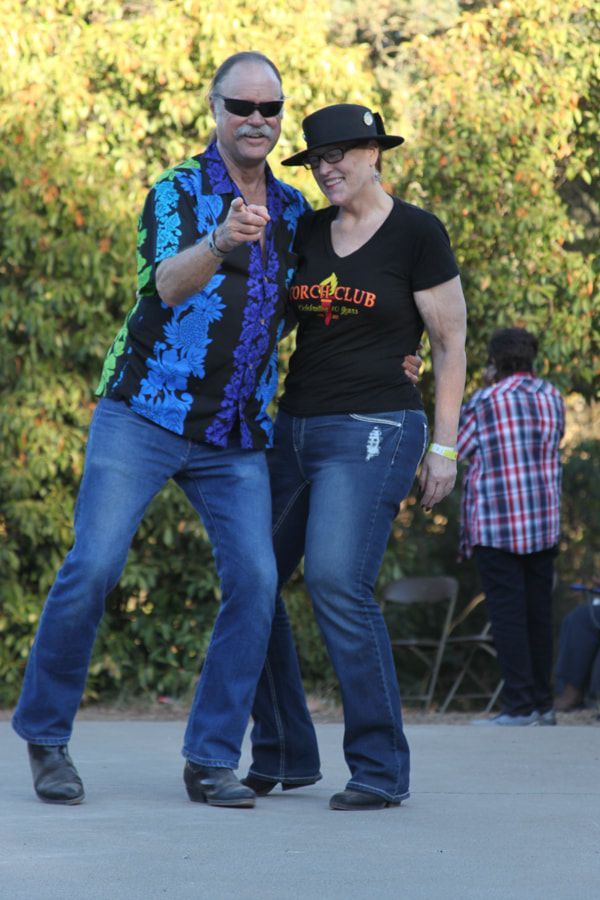 Two people dancing outdoors, one in sunglasses and a blue shirt, the other in a black hat and T-shirt.