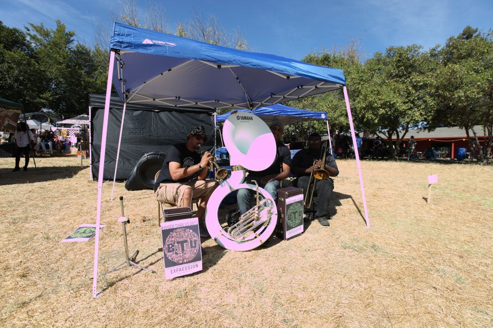 People sitting under a blue tent at an outdoor event, with a pink sign in front.