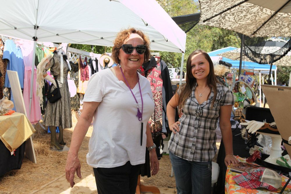 Two women smiling at an outdoor market stall under parasols, with colorful clothes and goods around them