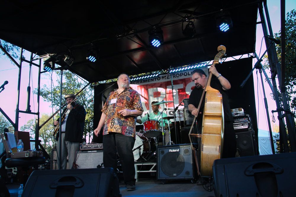 Musicians performing on an outdoor stage at dusk, with saxophone, guitar, and upright bass under colorful lights