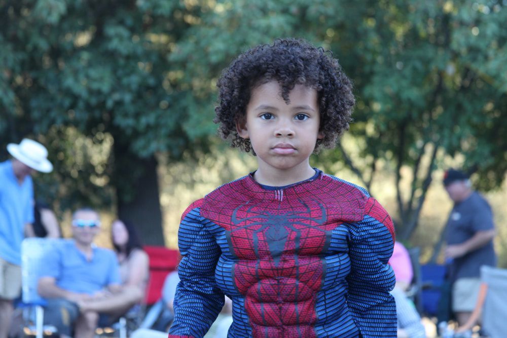 Child in a Spider-Man costume standing outdoors in a park, with people seated in the background.