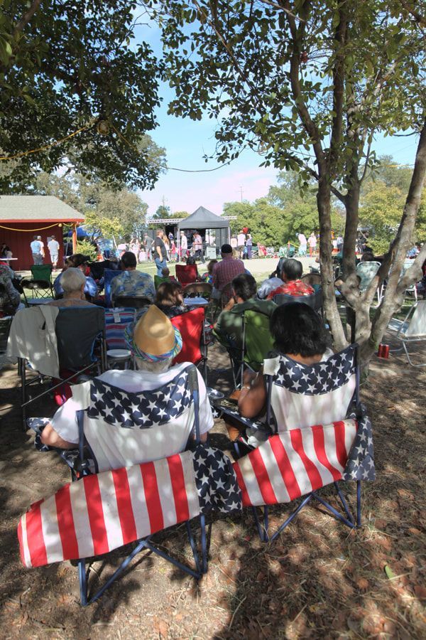 People seated in striped lawn chairs under trees, watching an outdoor event near a small stage.