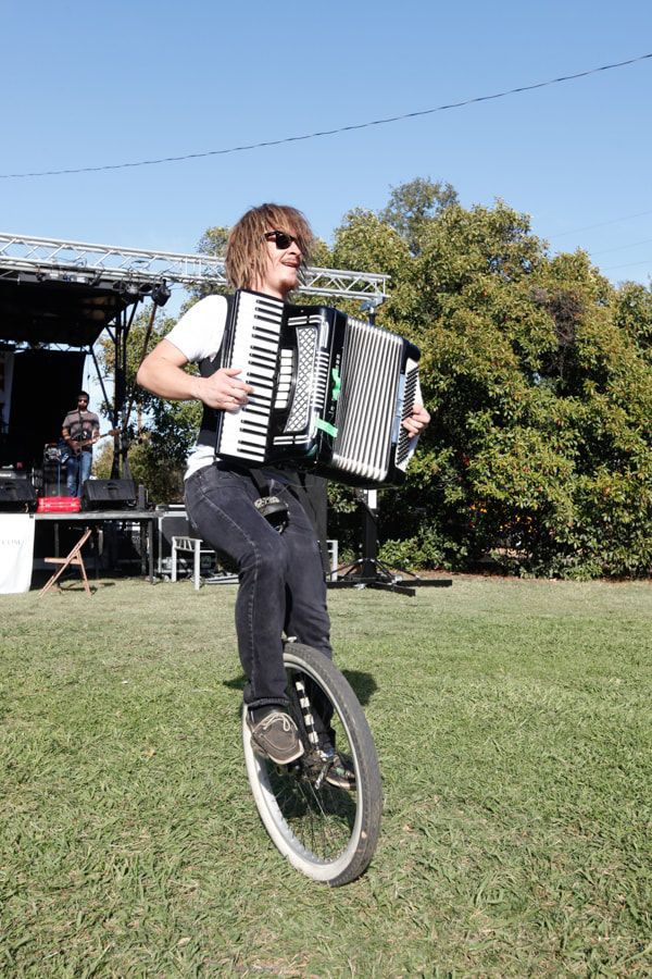 Person playing accordion while riding a unicycle on grass outdoors near a stage