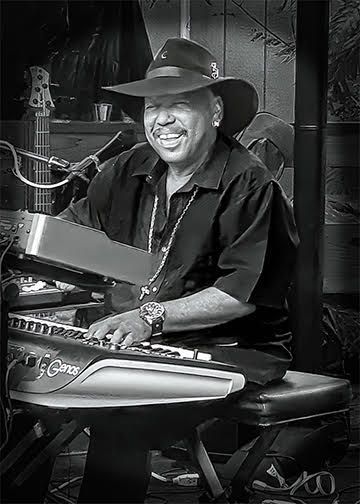 Smiling musician in a hat playing keyboard and guitar on stage, in black and white