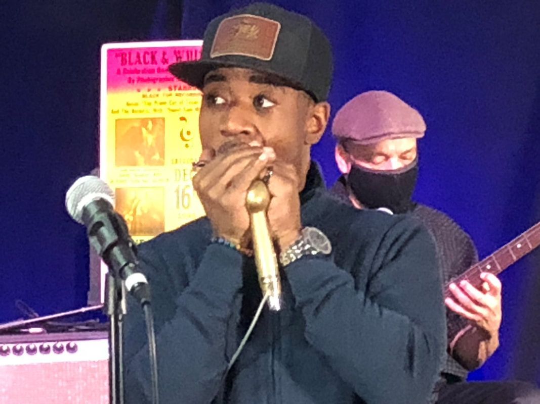 Musician onstage singing into a microphone, with another performer playing guitar in the background.