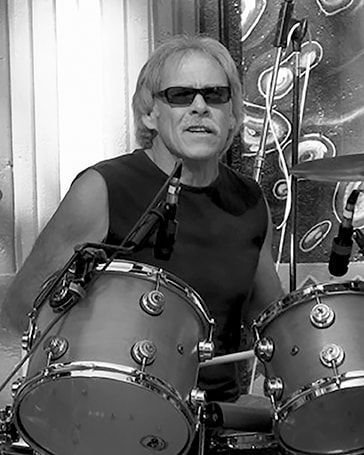 Drummer wearing sunglasses playing a drum kit onstage, with a microphone and abstract backdrop