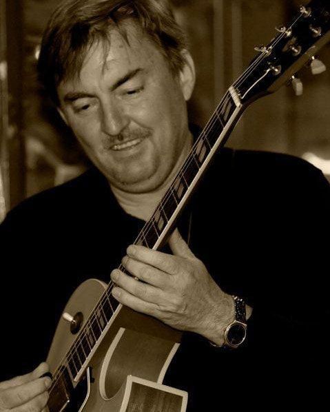 Man playing an electric guitar with eyes closed in a warm, sepia-toned close-up.