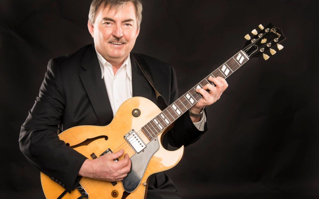 Man in a black suit holding a yellow electric guitar against a dark background