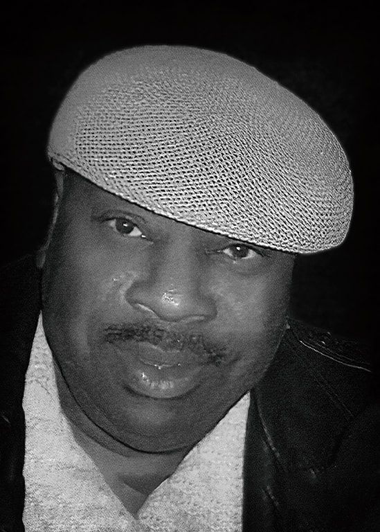 Black-and-white portrait of a person wearing a patterned cap, looking at the camera with a slight smile.