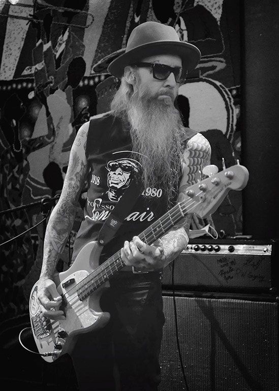 Bearded musician in sunglasses and hat playing bass guitar onstage in black-and-white.