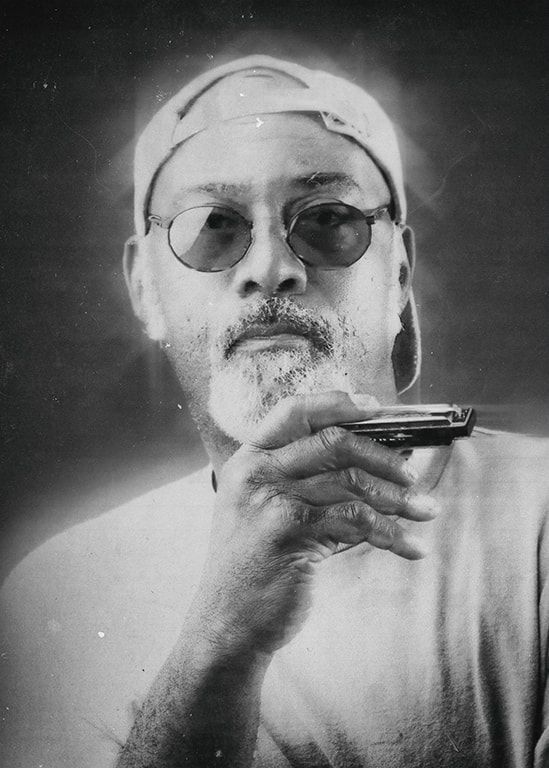 Black-and-white portrait of a man wearing glasses and a cap, holding a harmonica near his mouth