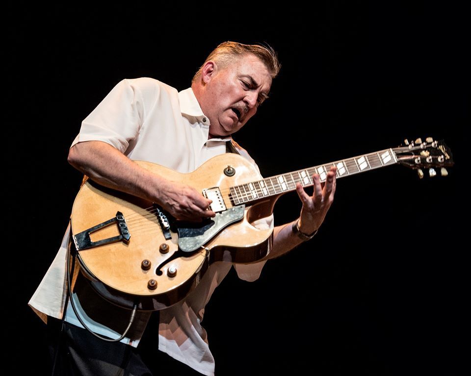 Man playing a tan electric guitar onstage against a black background, leaning into the instrument