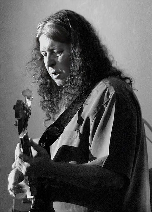 Black-and-white photo of a person playing an electric guitar, eyes closed, with long curly hair.
