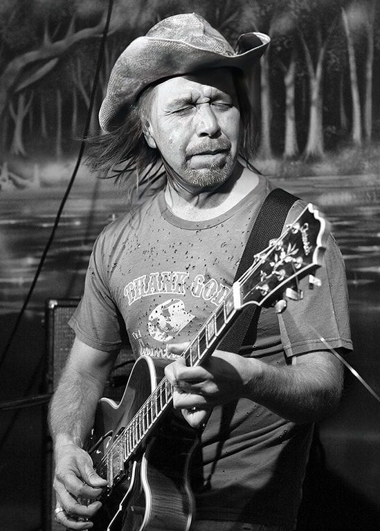 Man playing electric guitar outdoors, wearing a cowboy hat and T-shirt, looking to the side.