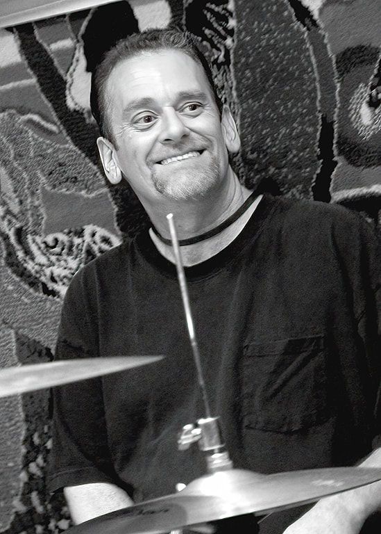 Man smiling behind a drum kit, holding a drumstick, in front of a patterned backdrop.