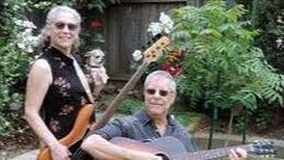 Two musicians with guitars outdoors, smiling in a leafy garden setting