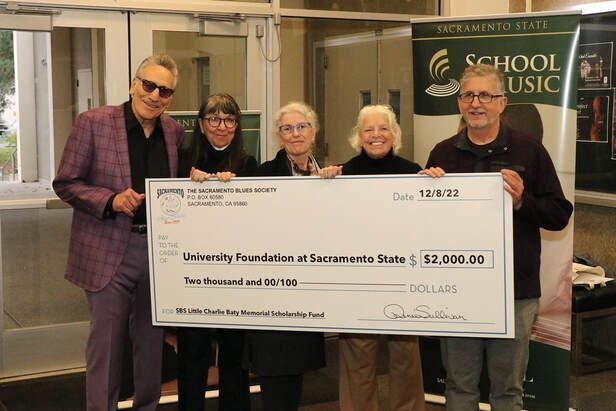 Four people hold a large donation check in front of a School of Music sign.