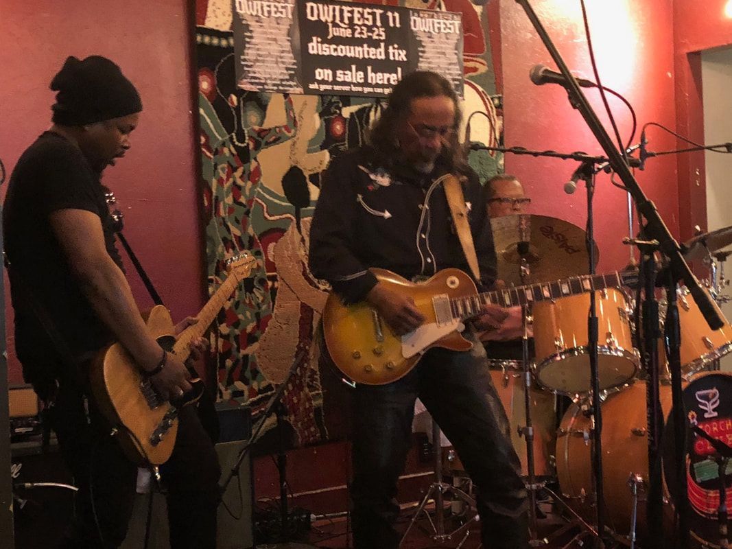 Two musicians playing electric guitars on a small stage with microphones and a drum kit in the background