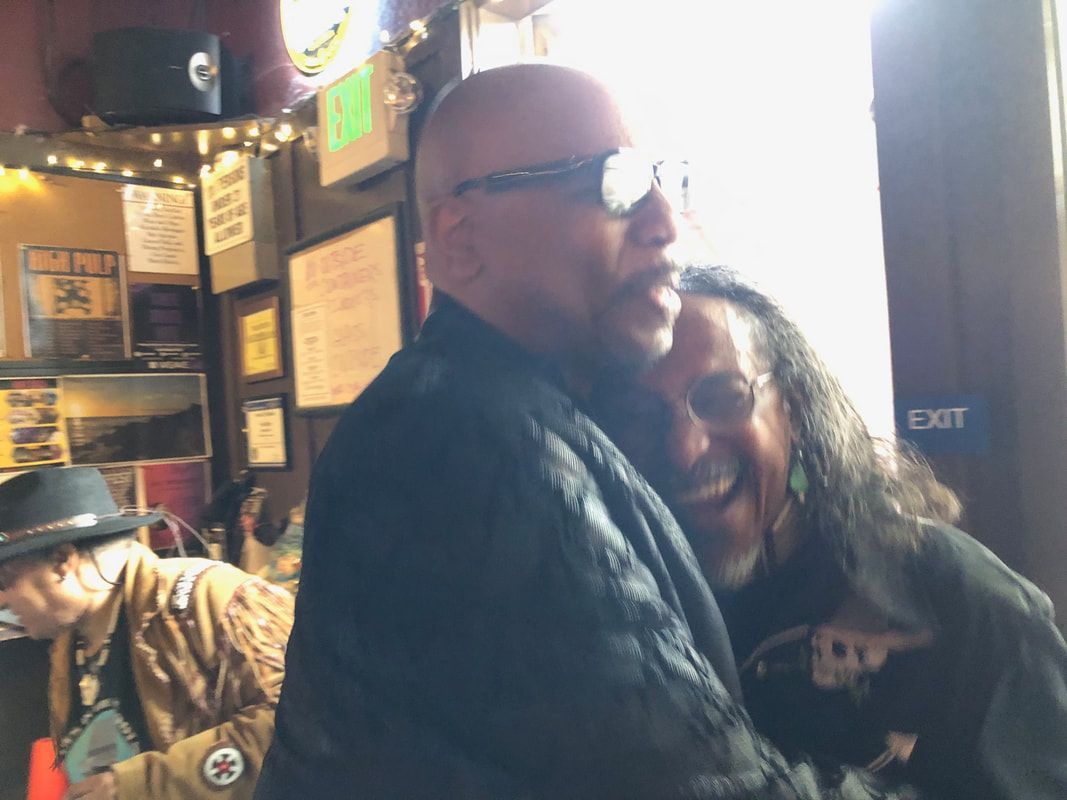 Two people hugging in a dimly lit bar, with one wearing glasses and the other smiling over his shoulder.