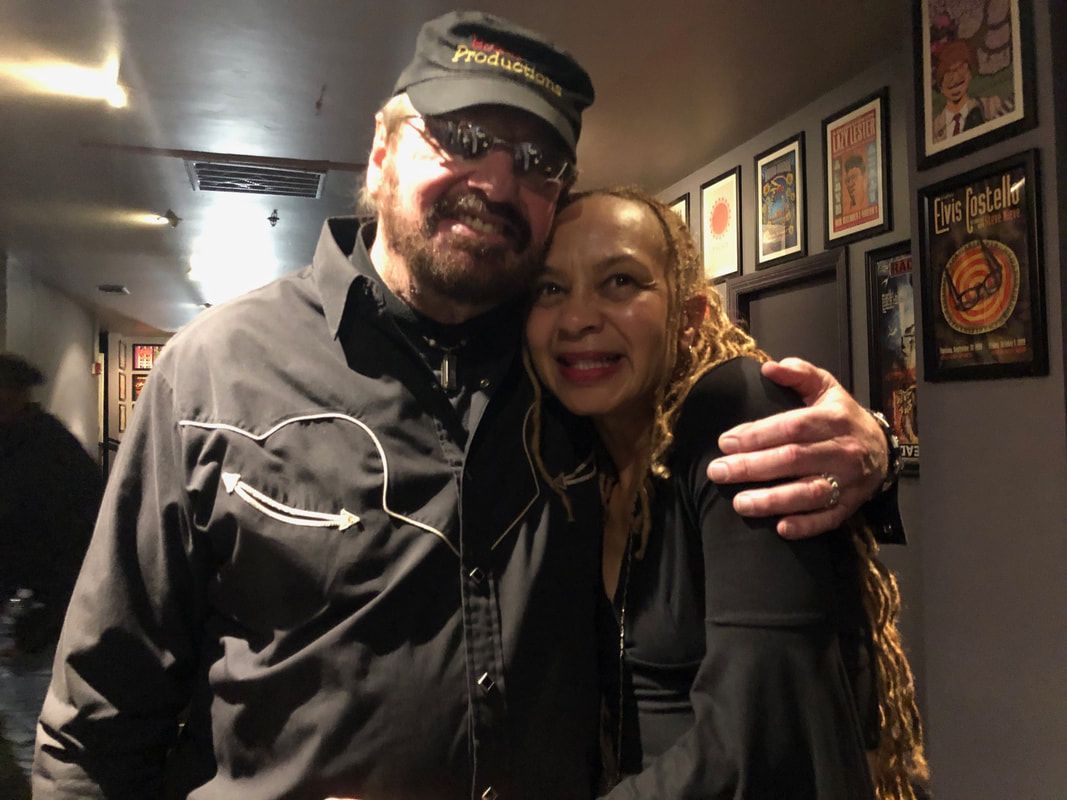 Two people smiling and hugging in a dim hallway with framed posters on the wall.