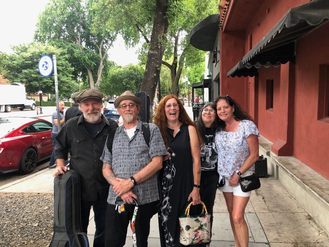 Four people smiling on a sidewalk beside a red building and parked cars under trees.