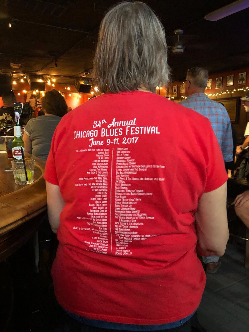 Person in red T-shirt with event schedule printed on the back, standing in a dimly lit bar.