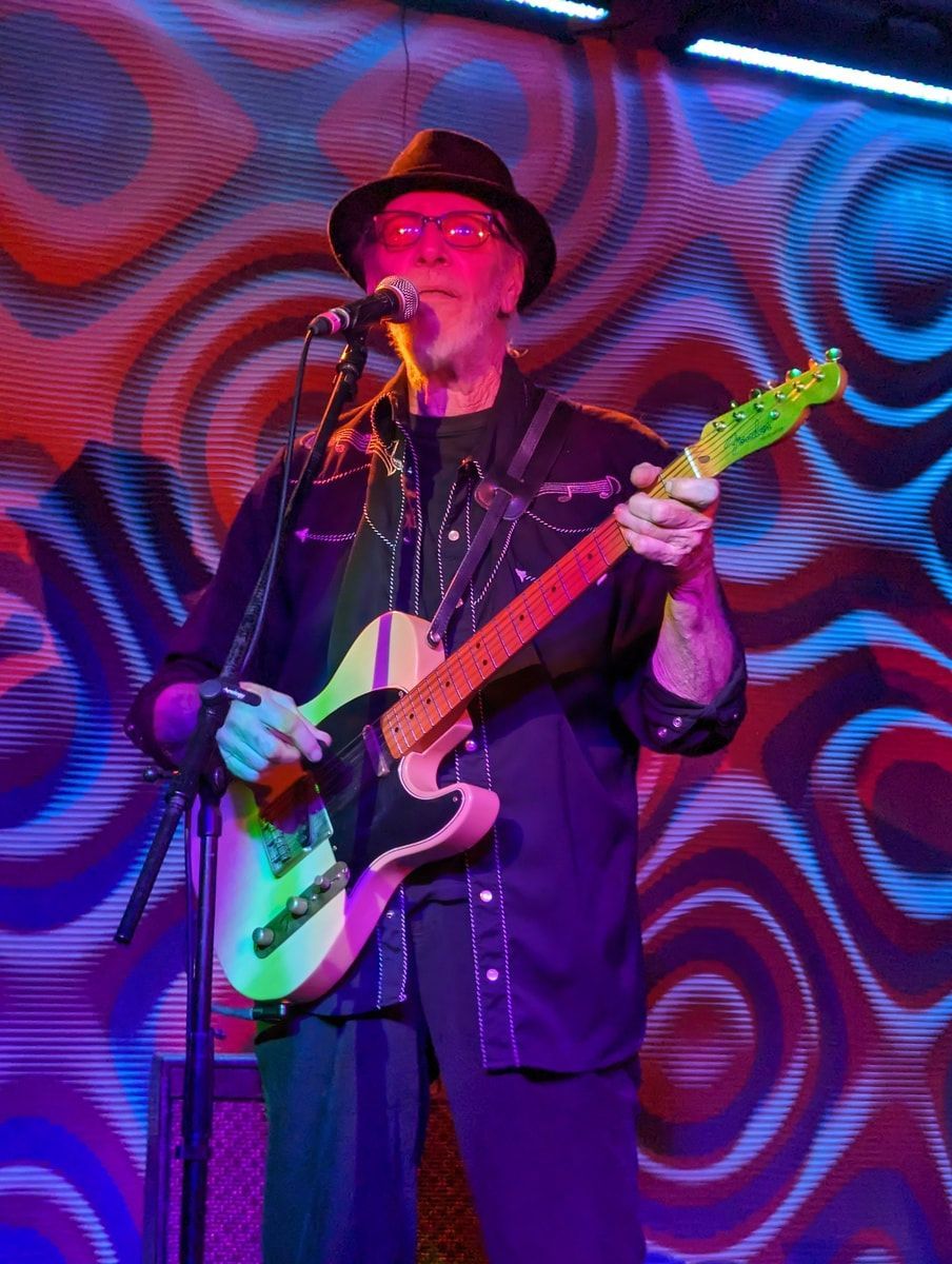 Musician in a black hat playing electric guitar under purple and red stage lights.