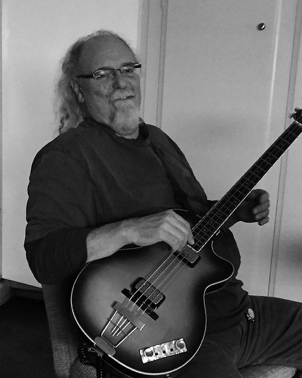Man seated indoors holding a sunburst bass guitar, wearing glasses and a dark shirt.