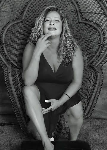 Woman in a black dress seated on a wicker chair, posing with a slight smile.