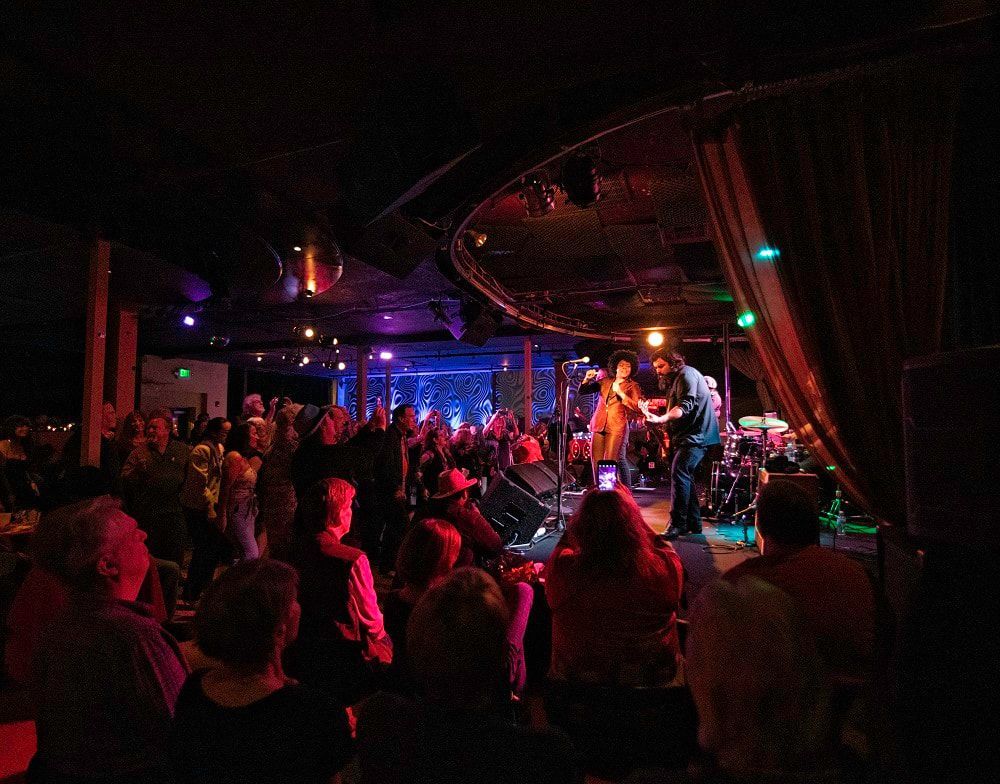 Crowded concert venue with stage lights, a live band, and audience facing the performance