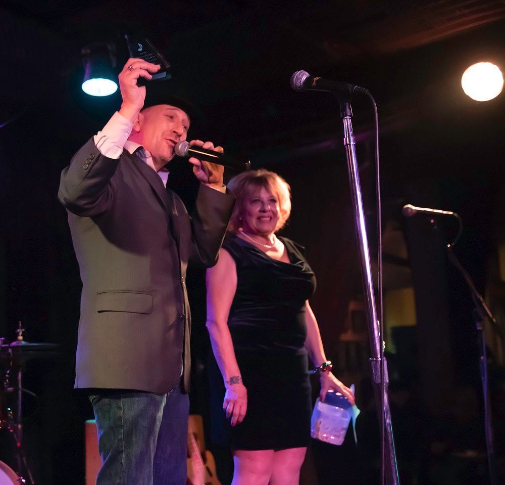 Two presenters on a stage, one speaking into a microphone under purple lights