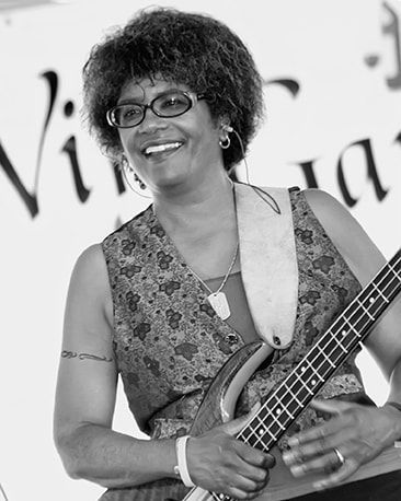 Smiling musician holding a bass guitar, wearing glasses and a patterned dress