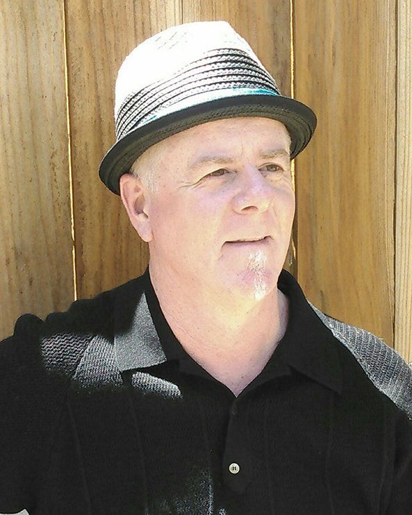 Man in a black shirt and striped hat standing against a wooden wall in bright sunlight