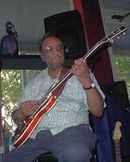 Musician seated indoors, holding a red electric guitar and playing on stage