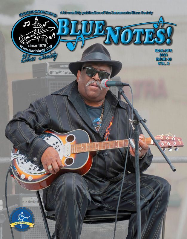 Musician in black hat and leather jacket plays guitar and sings into a microphone onstage, Blue Notes cover.