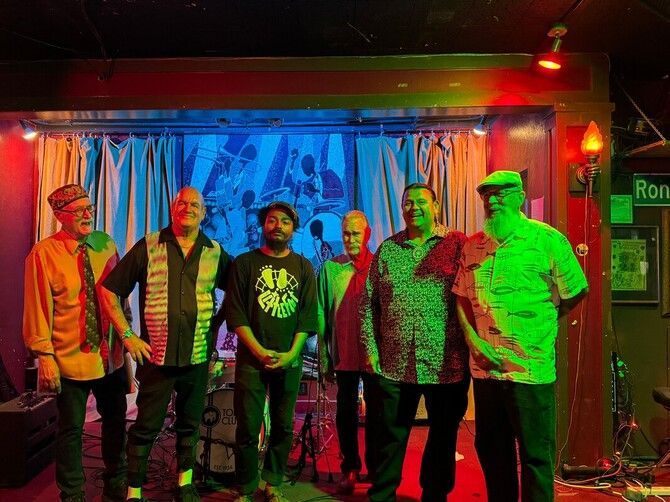 Six people stand on a small stage under colorful red and green lights in a dim bar.