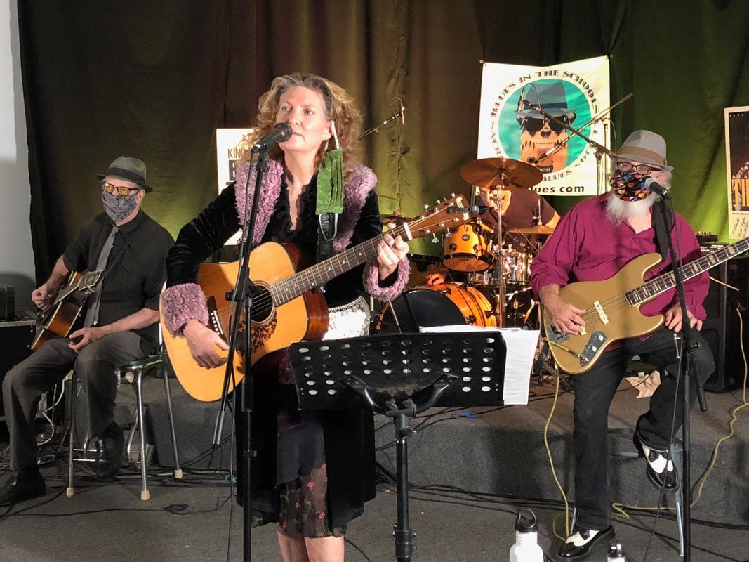 Band performing on stage, with singer at center, guitarists, drums, and music stands.