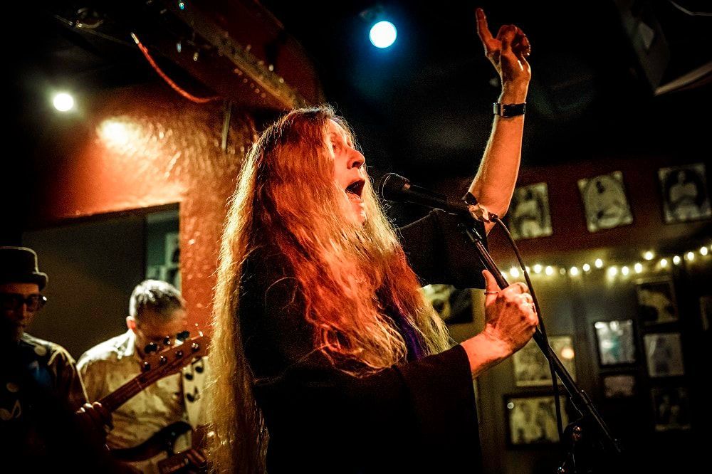 Singer with long red hair performing onstage under warm lights, microphone in hand, band visible in background