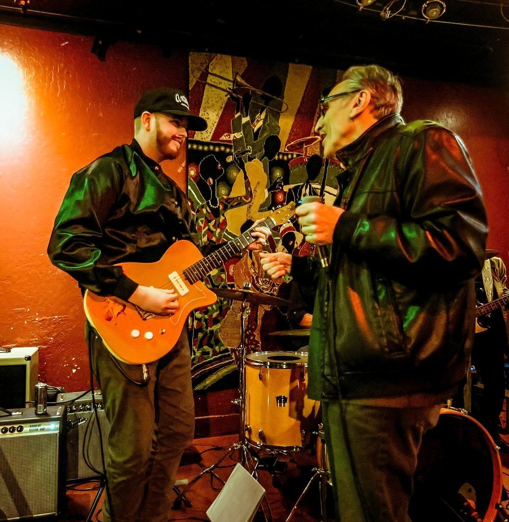 Two musicians performing live on stage, one playing an orange guitar under warm red lighting.