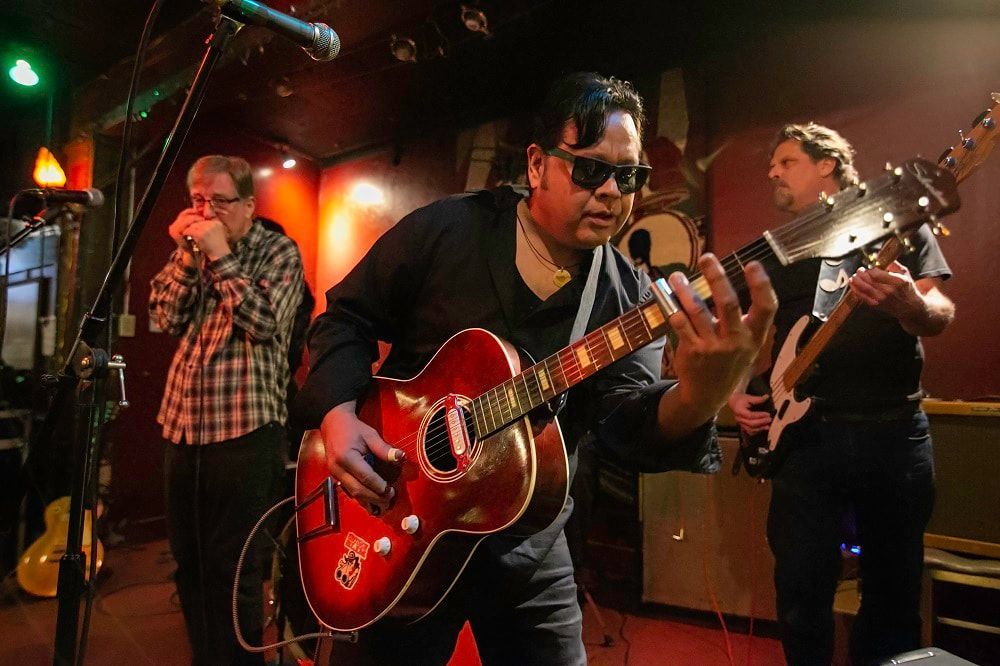 Live band performing on stage, with a man in sunglasses playing a red guitar in the foreground.
