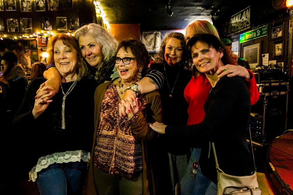 Five friends smiling and posing together in a warmly lit bar or pub.