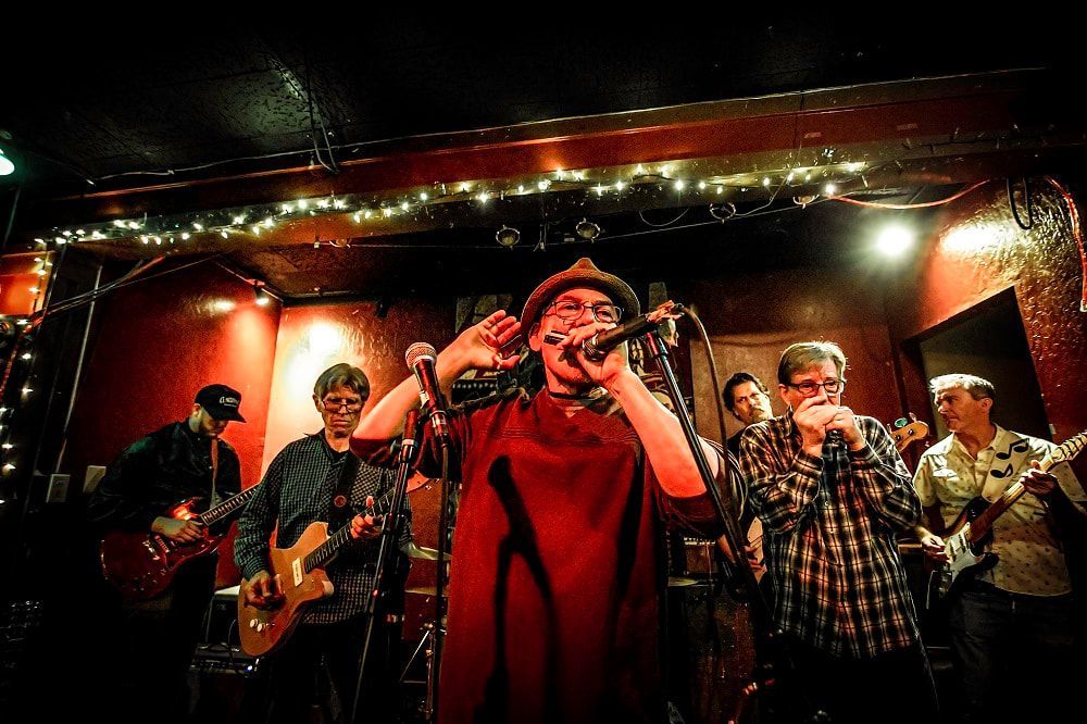 Live band performing on a dimly lit stage, with a singer in a red coat at center microphone.