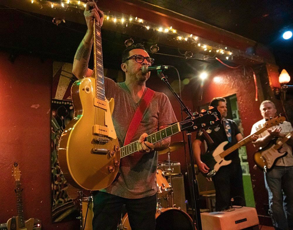 Singer-guitarist on stage raises a yellow guitar in a dimly lit club with bandmates behind him