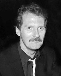Black-and-white portrait of a man in a dark suit with a mustache, facing the camera and smiling slightly